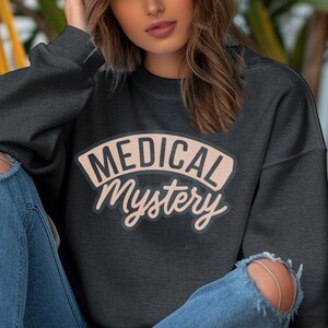 以下が含まれることがあります： レトロなフォントで「MEDICAL Mystery」と書かれたピーチ色のパッチが付いたダークグレーのスウェットシャツ。モデルは青いジーンズとグレーのベースボールキャップを着用しています。