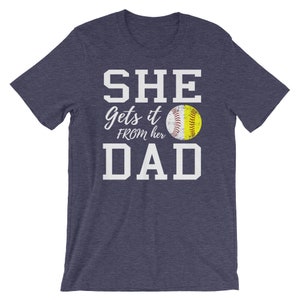 May include: A navy blue t-shirt with white text that reads "SHE gets it FROM her DAD" and a yellow softball graphic.