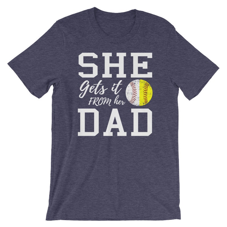 May include: A navy blue t-shirt with white text that reads "SHE gets it FROM her DAD" and a yellow softball graphic.