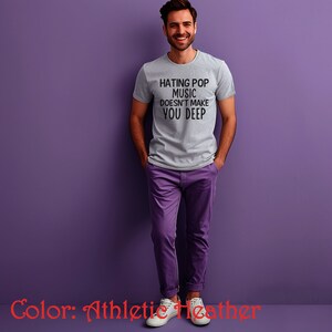 Puede incluir: Una camiseta gris con el texto "Hating Pop Music Doesn't Make You Deep" impreso en la parte delantera. La camiseta est&aacute; siendo modelada por una persona sobre un fondo morado.