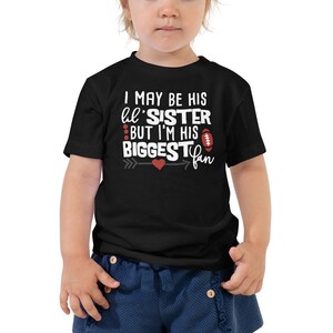 May include: Black toddler t-shirt with white text that reads "I MAY BE HIS lil' SISTER BUT I'M HIS BIGGEST fan". The design includes a red heart and a football graphic. The shirt is a short-sleeved crew neck.