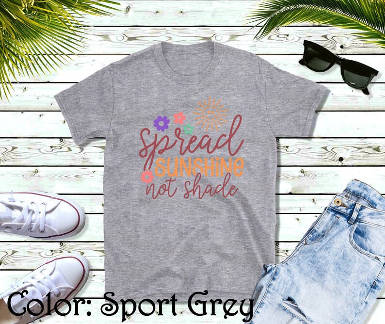 May include: A heather grey t-shirt with the text "Spread Sunshine not shade" in orange and brown lettering. The text is surrounded by small flowers and a sun graphic.