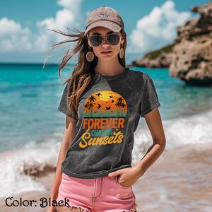 Puede incluir: Una camiseta negra con un aspecto desgastado que presenta un gráfico de puesta de sol con palmeras y pájaros. El texto "FOREVER CHASING SUNSETS" está impreso en una fuente de estilo vintage.