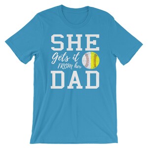 May include: A light blue t-shirt with white text that reads "SHE gets it FROM her DAD" and a softball graphic.
