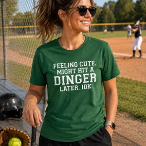May include: A dark green short-sleeved t-shirt featuring white block lettering that reads: FEELING CUTE. MIGHT HIT A DINGER LATER. IDK. The shirt has a crew neckline and a standard fit, constructed from a solid-colored fabric with a smooth texture.