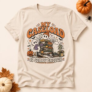 Pode incluir: Uma t-shirt de manga curta cor de creme com um gráfico de três fantasmas a trabalhar numa secretária cheia de papéis. O texto diz MY CASELOAD IS SCARY ENOUGH. O design inclui abóboras, uma caneca de café e teias de aranha.