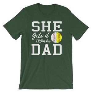 May include: Green short-sleeved t-shirt with white text that reads "SHE gets it FROM her DAD" and a yellow softball graphic.