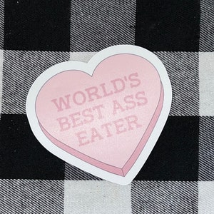 Puede incluir: Una pegatina rosa en forma de corazón con el texto "WORLD'S BEST ASS EATER" impreso en negro.