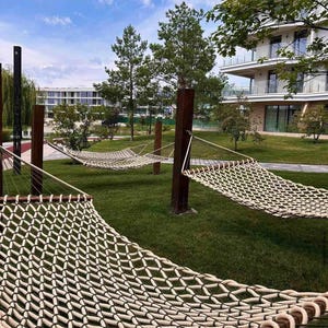 Peut inclure: Plusieurs hamacs en corde tressée sont tendus entre des poteaux en bois dans un espace herbeux. Les hamacs sont beiges et bruns, avec des arbres, des bâtiments et un ciel bleu nuageux en arrière-plan. La scène évoque la détente et les loisirs.