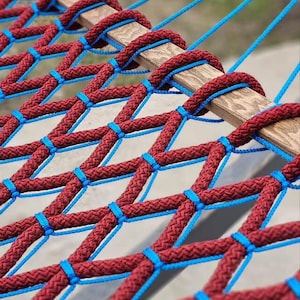 May include: Close-up of a hammock with a woven design. The hammock features a burgundy rope netting, accented with blue rope ties. The wooden support beams are visible, and blue ropes extend from the top.