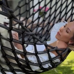 May include: A person relaxing in a black rope hammock outdoors. The hammock is suspended between two supports. A white dog is resting in the hammock with the person. The person is wearing a white shirt and is lying down on the hammock.