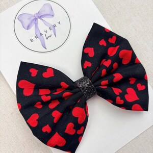 May include: A black fabric bow with a pattern of red hearts. The bow has a glittery black center and is set against a white background. The logo "Bravery Bow" is visible in the upper left corner.