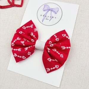 May include: A red fabric bow with a white arrow and heart pattern, attached to a white card. The bow has a white center and is labeled with the text "Bravery Bow". Decorative red glitter hearts are in the background.