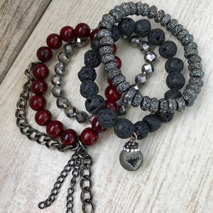 Pode incluir: Um conjunto de pulseiras de contas em tons de vermelho, cinza e preto. Uma pulseira apresenta um detalhe de corrente, enquanto outra tem um pingente com um design em forma de coração. As pulseiras são feitas com contas e pedras de lava.