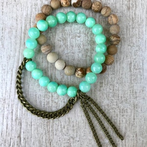 Op de afbeelding: Drie armbanden met verschillende kralenstijlen. Eén armband heeft lichtgroene, ronde kralen. Een andere heeft bruine en bruingespikkelde kralen. Een derde armband heeft een ketting en kwastjes in brons.