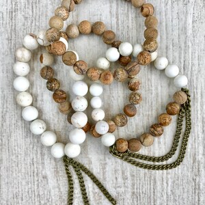 Pode incluir: Três pulseiras de contas. Duas pulseiras apresentam contas redondas, castanhas claras e bege. A terceira pulseira tem contas brancas. Todas as três pulseiras têm um detalhe de corrente dourada pendurado na parte inferior.