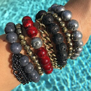 Könnte beinhalten: Eine Sammlung von Armbändern mit Perlen in verschiedenen Stilen und Farben. Die Armbänder haben graue, rote, schwarze und metallische Perlen sowie eine goldfarbene Kette und facettierte Perlen. Die Armbänder sind gestapelt und zeigen eine Vielzahl von Texturen und Designs.