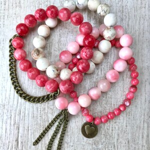 Könnte beinhalten: Drei Armbänder mit rosa, roten und weißen Perlen. Eines hat eine Kette und Quaste, ein anderes einen Herzanhänger. Die Perlen variieren in Größe und Textur und erzeugen einen geschichteten Look.