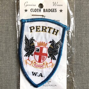 May include: A white woven cloth badge with blue edging. The badge features the text "PERTH" in gold lettering, a coat of arms with two black birds, and the text "W.A. AUSTRALIA". The badge is packaged in a clear plastic bag with a white card that reads "Genuine Woven Cloth Badges by ...tions".