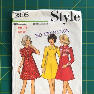 May include: A vintage sewing pattern labelled "Style" with illustrations of three dresses. The pattern includes sizing information and the text "NO EXCHANGE". The dresses are in red, yellow, and red with a gingham pattern.