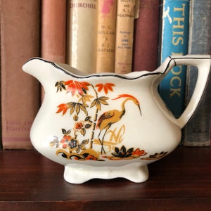 May include: A small, white ceramic creamer with a black rim and handle. The creamer features a decorative design of a crane, bamboo, and flowers in orange, gold, and black. The creamer is displayed on a dark wooden surface.