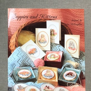 May include: A craft book titled "Puppies and Kittens" with cross-stitch designs. The image shows framed cross-stitch pieces and round boxes with embroidered animal designs. A pink ball of yarn and a blue crocheted blanket are also visible. The designs are by Julie McLaughlin.