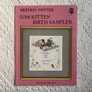 May include: A pink and white cross stitch pattern featuring Beatrix Potter's Tom Kitten. The pattern includes a framed image of two kittens playing with a butterfly, surrounded by flowers and vines. The text below the image reads "christopher ryan cooke april 5, 1994 7lbs. 7oz."