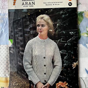 May include: A knitting pattern for a cardigan, featuring a woman wearing a light grey cardigan with a cable knit design. The pattern is for sizes 34/36 and 36/38 inch bust and requires 28 (30) ounces of yarn. The pattern is titled "sunbeam ARAN OILED YARN" and is priced at 9d.
