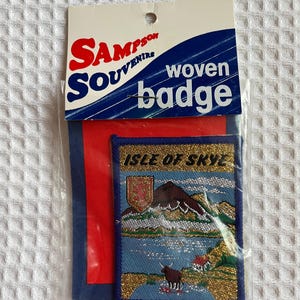 May include: A woven badge souvenir from Sampson Souvenirs, depicting the Isle of Skye. The rectangular badge has a blue border and gold detailing, packaged with a red and blue backing. The packaging reads 'woven badge'.