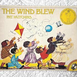 May include: A children's book cover for "The Wind Blew" by Pat Hutchins. The illustration shows people reaching for items caught in the wind, such as a kite, hat, and balloon. The cover has a yellow and white background with the text in yellow.