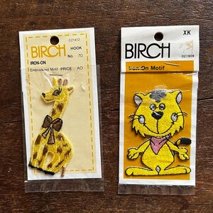May include: Two iron-on patches, one featuring a yellow giraffe with a brown bow and the other a yellow cat with a pink bandana. Both patches have the word "BIRCH" printed on the packaging.
