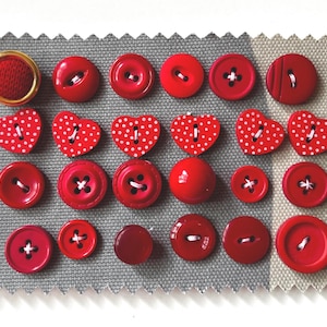 May include: A selection of red buttons in different shapes and sizes, including heart-shaped and round. Some have white polka dots or white thread. The buttons are displayed on a grey fabric swatch.