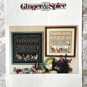 May include: A printed sampler pattern by Ginger & Spice, featuring two framed cross-stitch samplers with alphabet and floral designs. The text includes the words "Stitchers Sampler" and "Charted Designs". Various sewing notions are arranged below the framed samplers.