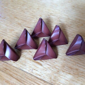 May include: Six triangular, burgundy-coloured buttons with a glossy finish are arranged on a light brown wooden surface. The buttons have a geometric design, with each side of the triangle slightly curved.