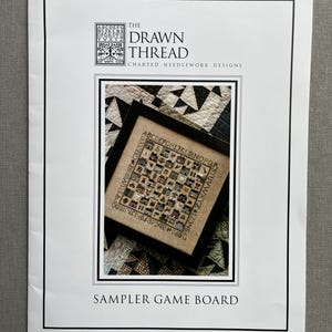 May include: A printed paper with the text "THE DRAWN THREAD CHARTED NEEDLEWORK DESIGNS" and "SAMPLER GAME BOARD". The image features a framed cross-stitch sampler with a house design. The sampler is in a black frame.