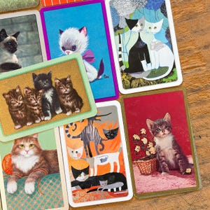 May include: A collection of rectangular playing cards featuring various cat illustrations, including a group of four kittens on a gold background, a tabby cat with a yarn ball, and stylized artistic depictions of cats in vibrant colors.