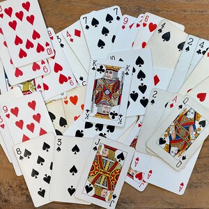 May include: A scattered assortment of playing cards featuring suits of hearts, spades, diamonds, and clubs. The cards display various numbers and face cards, including kings, queens, and jacks. The cards are on a wooden surface.
