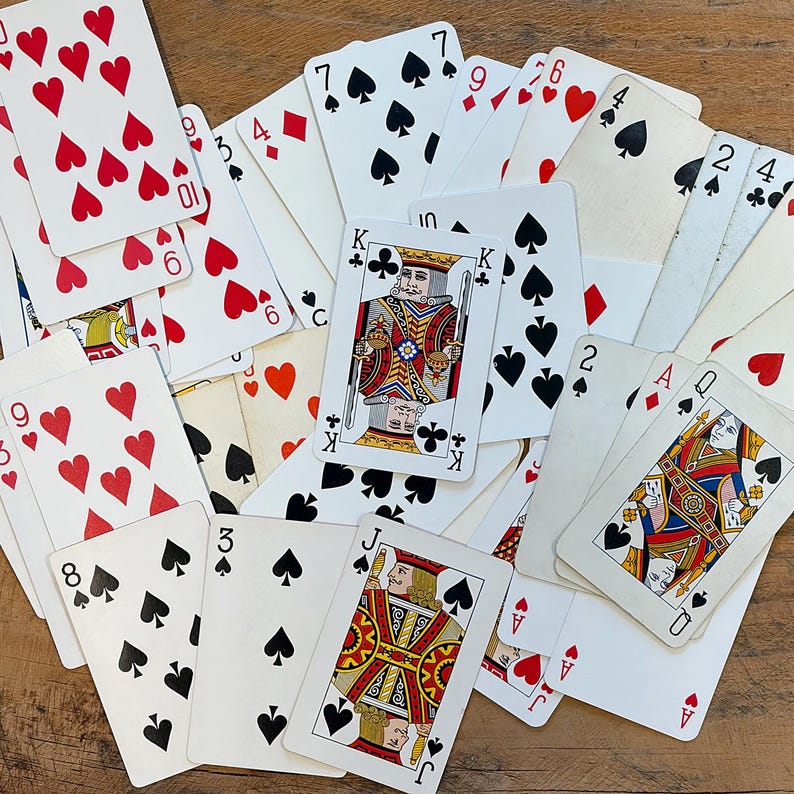 May include: A scattered assortment of playing cards featuring suits of hearts, spades, diamonds, and clubs. The cards display various numbers and face cards, including kings, queens, and jacks. The cards are on a wooden surface.