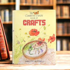 May include: The Country Diary Book of Crafts by Annette Mitchell, a book cover with a yellow background and a floral design featuring red flowers and a butterfly. The book title is in red text and the author's name is in black text.