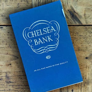 May include: A blue book with the words "CHELSEA BANK" in white script. The text "AN ALL RAG PAPER OF FINE QUALITY" is printed at the bottom. The book is on a wooden surface.
