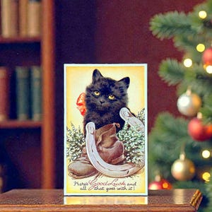 Könnte beinhalten: Vintage Postkarte mit einer schwarzen Katze mit gelben Augen und einer roten Schleife. Sie zeigt ein Hufeisen und einen braunen Stiefel. Der Text am unteren Rand der Karte lautet: "Here's Good Luck and all that goes with it!"