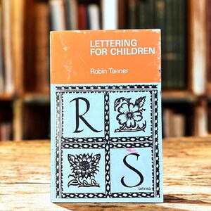 May include: A vintage book titled "Lettering for Children" by Robin Tanner. The cover has an orange top and a light blue section with the letters "R" and "S" and floral illustrations. The book is set against a blurred background of bookshelves.