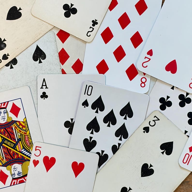 May include: A collection of scattered playing cards featuring various suits including spades, hearts, diamonds, and clubs. The cards display numbers, an ace, and a king, with red and black ink on white cardstock, showing symbols and numerical values.
