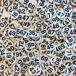 Könnte beinhalten: Draufsicht auf eine Sammlung von cremefarbenen, runden Spielmarken. Jede Marke trägt eine schwarze Nummer. Die Zahlen, von 1 bis 99, sind auf den Marken aufgedruckt, die auf einer hellen Oberfläche verstreut sind.