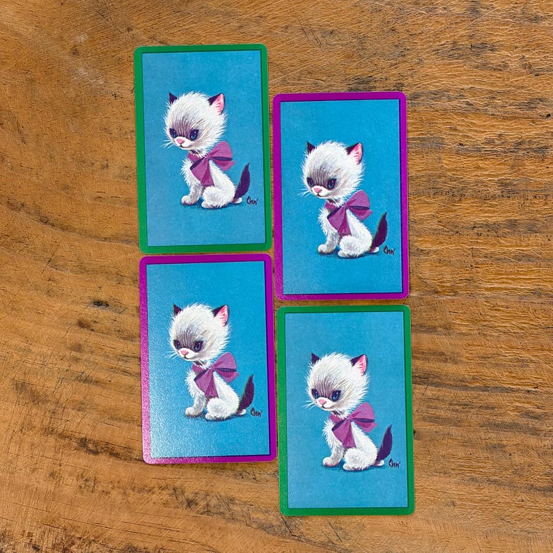 May include: Four vintage playing cards featuring a white kitten with gray fur, pink ears, and a purple bow. Each card has a blue background and a colored border: green or purple. The cards are arranged on a wooden surface.