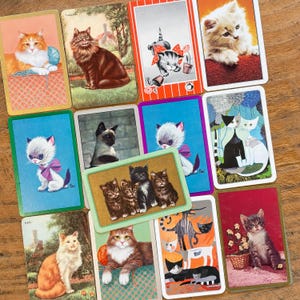May include: A collection of rectangular playing cards featuring various cat illustrations, including kittens and adult cats in diverse artistic styles, colors, and settings, arranged in a scattered layout on a wooden surface.