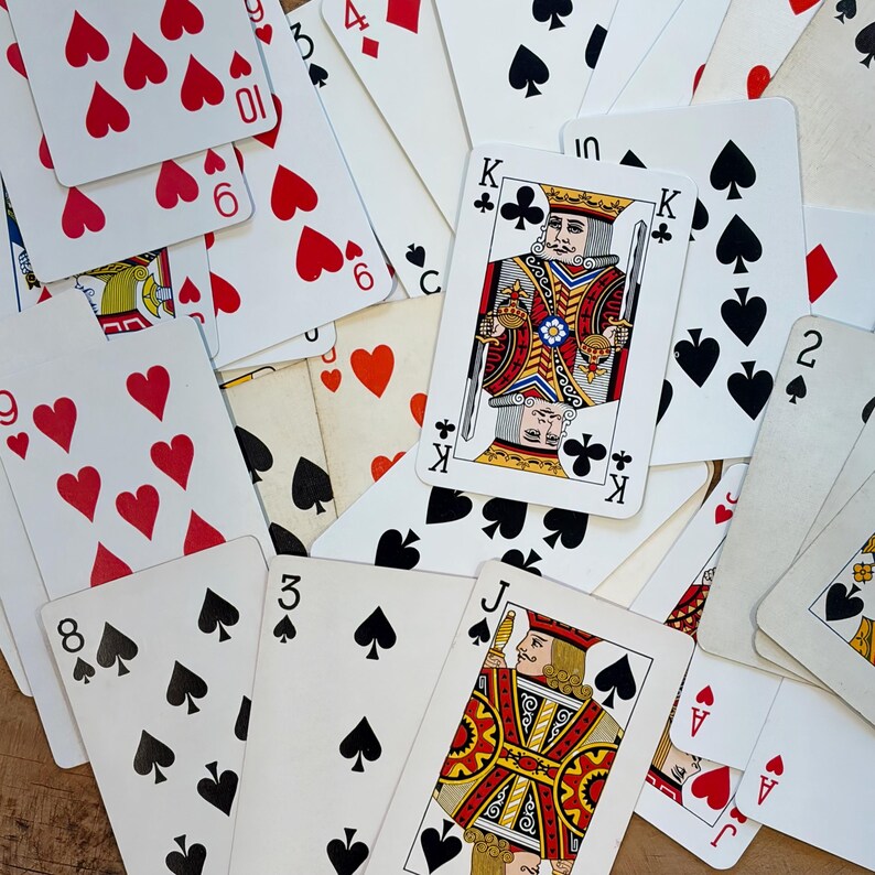 May include: A collection of playing cards scattered across a surface. The cards feature various suits, including hearts, spades, and clubs. A King card is prominently displayed, along with other numbered and face cards. The cards are white with red, black, and gold details.