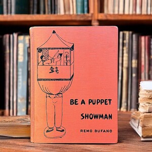 May include: A vintage book titled "Be a Puppet Showman" by Remo Bufano. The cover features a red background with a black illustration of a puppet show stage with two puppets inside. The book is sitting on a wooden surface.
