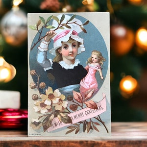 Ancienne carte de voeux de Noël victorienne représentant une jeune fille coiffée d'un bonnet avec sa poupée, une belle chromolithographie vintage Merry Xmas