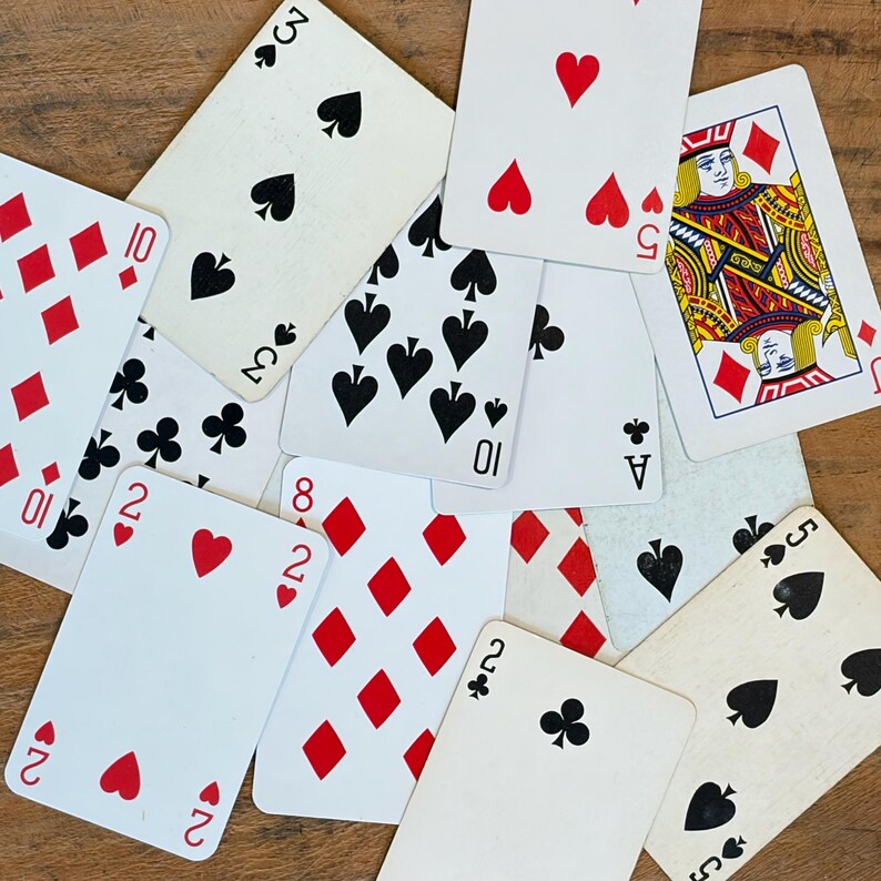 May include: A collection of scattered playing cards featuring various suits including hearts, diamonds, spades, and clubs. The cards display numerical values and a face card with a red and gold design. The cards are white with black and red ink.